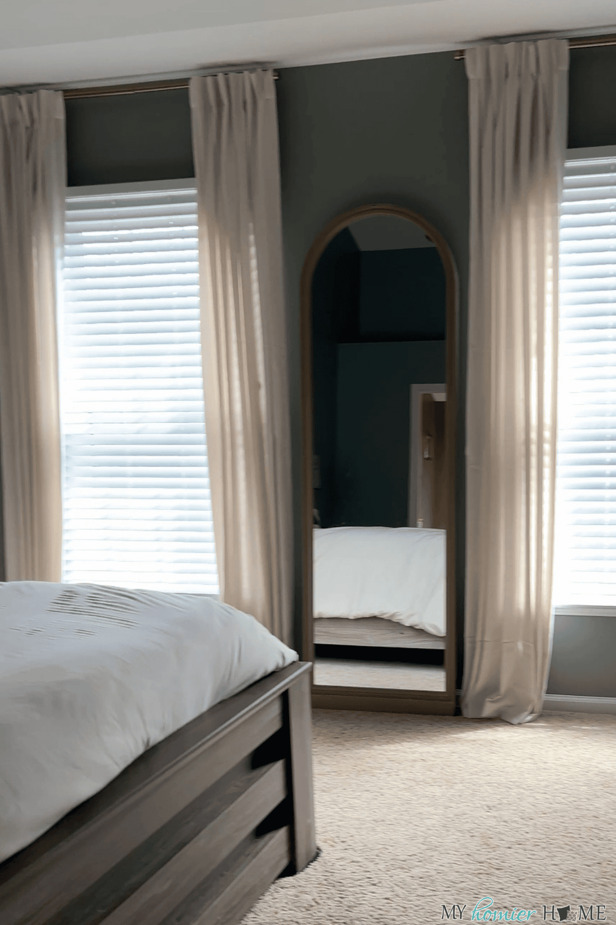 A bedroom with a wooden bed frame, two large windows with sheer curtains—perfect for those seeking bedroom curtain ideas—beige carpet, and a tall arched mirror reflecting part of the bed.