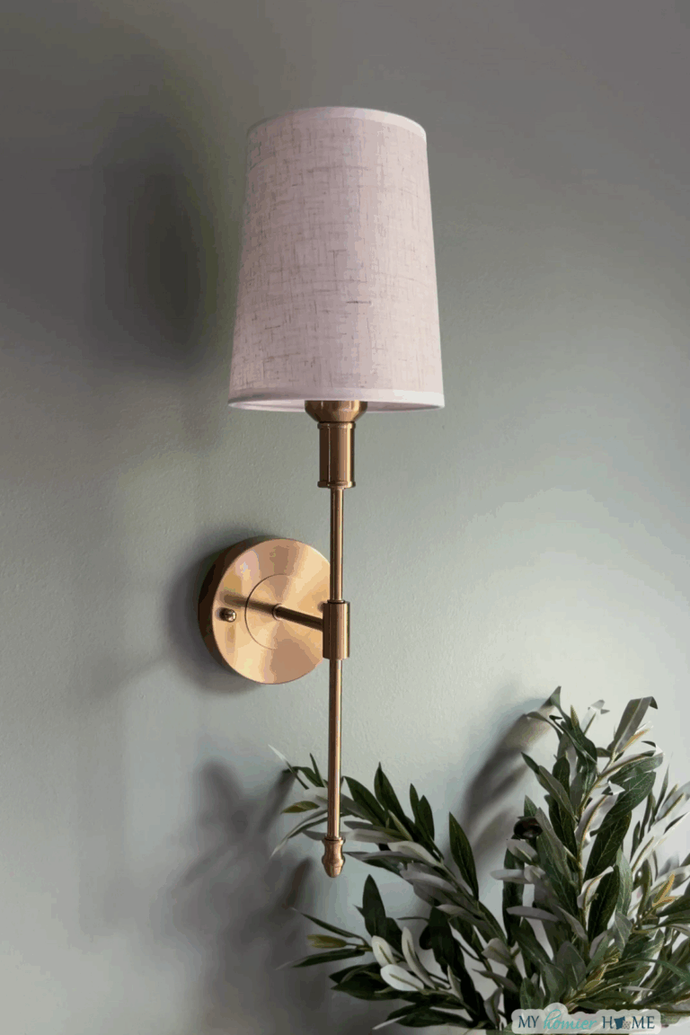 A gold wall sconce with a white fabric lampshade is mounted on a light green wall above green leafy branches—perfect for those seeking elegant bedroom lighting ideas.