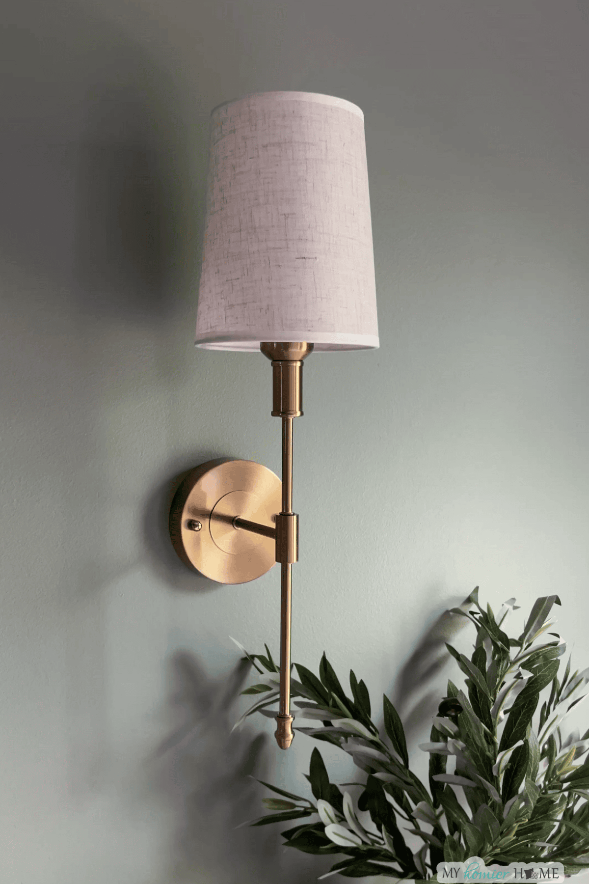 A gold wall sconce with a white fabric lampshade is mounted on a light green wall above green leafy branches—perfect for those seeking elegant bedroom lighting ideas.