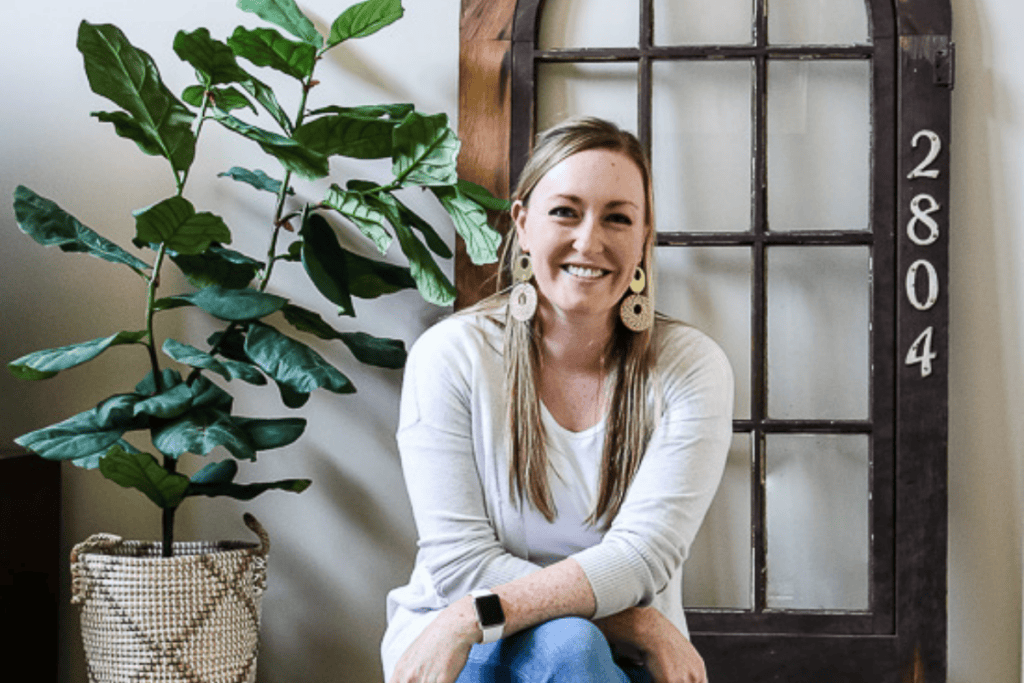 A woman, dressed in a white top and blue jeans, smiles as she sits by a potted plant next to a window marked with the numbers 2804. The scene exudes tranquility and charm, perfectly capturing a moment of simple joy amidst natural light.