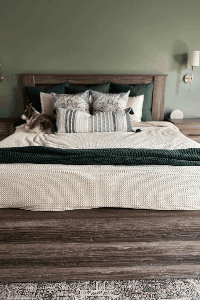 A neatly made bed with neutral and green bedding features multiple decorative pillows thoughtfully placed—showcasing how to arrange pillows on a king bed—with a cat lying on the left side and wooden headboard and side tables in view.