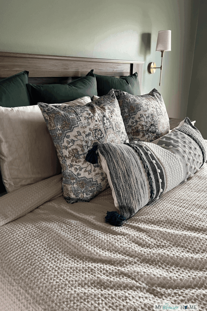 A neatly made bed with textured bedding, patterned throw pillows, dark green back pillows, and a wall-mounted reading light shows how to arrange pillows on a king bed for both comfort and style.