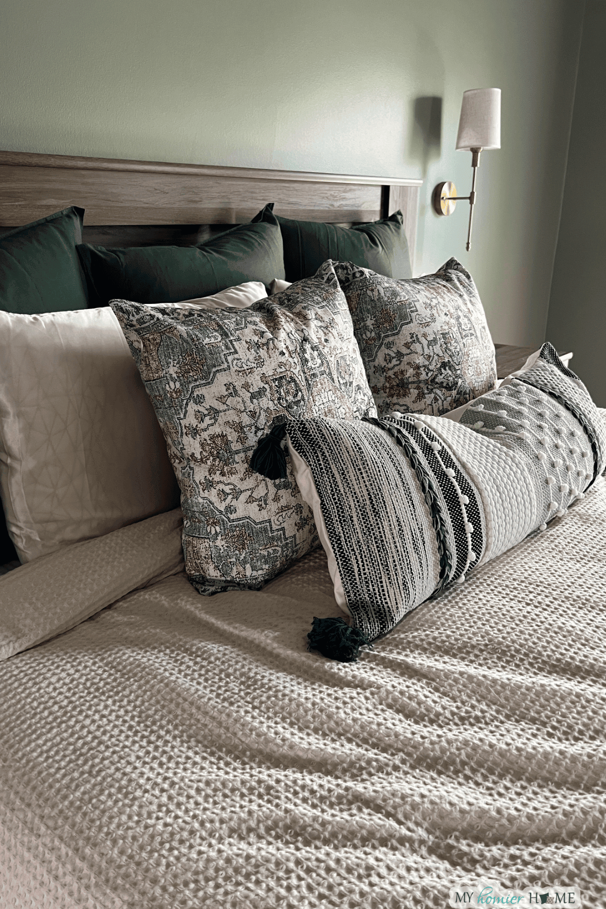 A neatly made bed with textured bedding, patterned throw pillows, dark green back pillows, and a wall-mounted reading light shows how to arrange pillows on a king bed for both comfort and style.