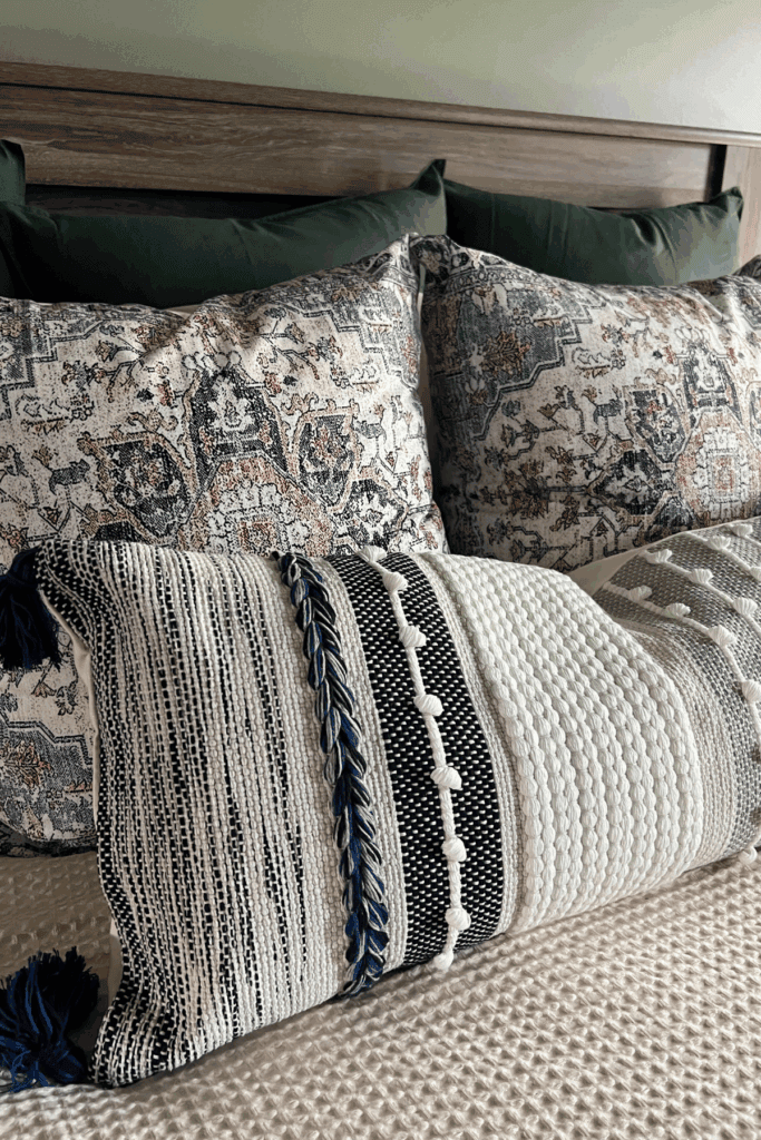 Close-up of a neatly made bed with patterned pillows in blue, white, and orange tones against a wooden headboard, offering inspiration for how to arrange pillows on a king bed. A textured decorative pillow is placed in front.