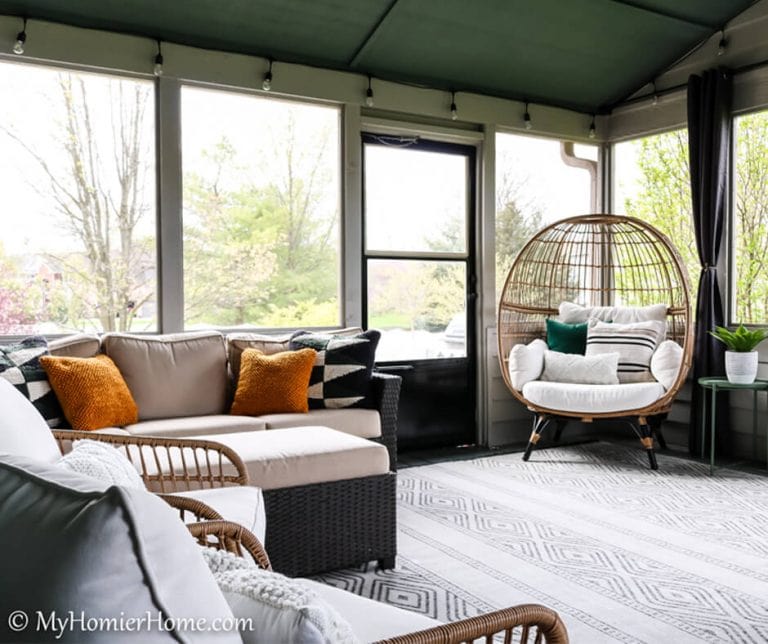 A cozy sunroom with a mix of wicker and cushioned furniture, including a hanging chair, and a patterned rug. A door opens to the outside, blending seamlessly with outdoor living room ideas. The room has large windows offering natural light and greenery views.