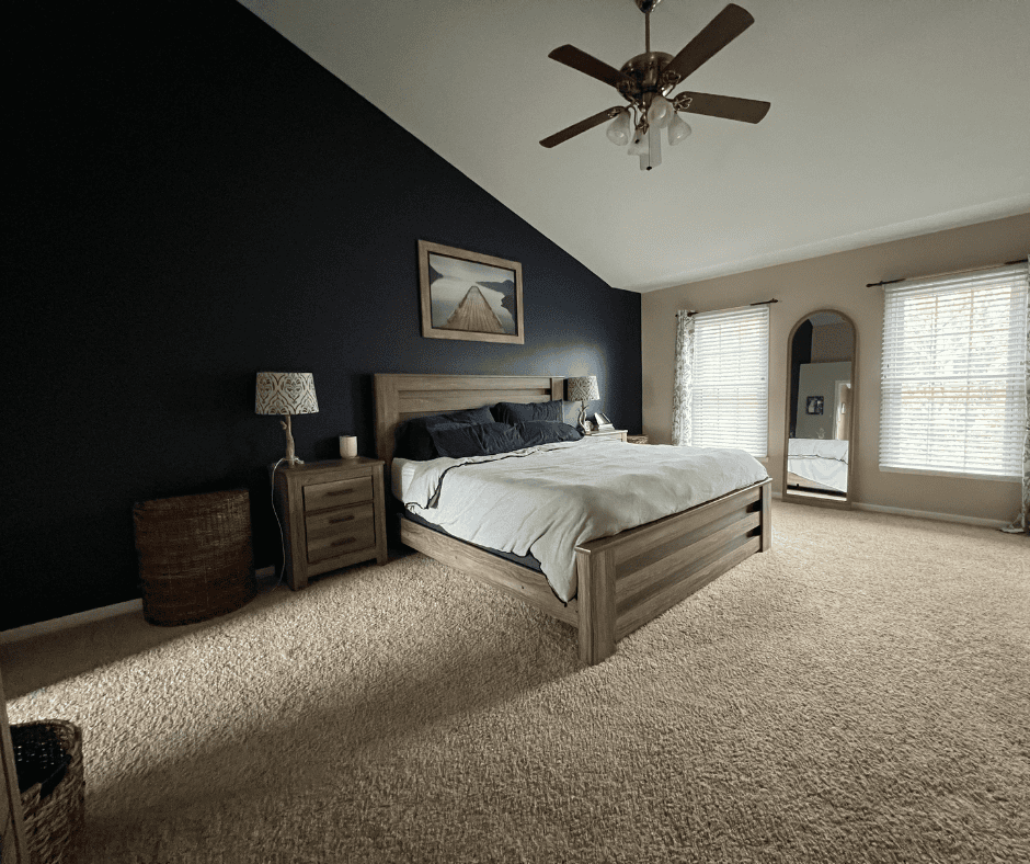 Primary bedroom ideas come to life in this space featuring a large bed, dark accent wall, and ceiling fan. It includes bedside tables, lamps, a wicker basket, and a framed wall picture. Natural light streams in through two windows for an airy ambiance.