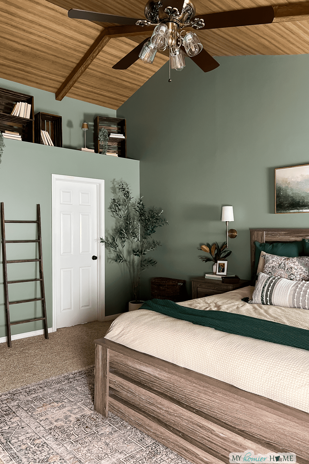 A primary bedroom makeover featuring a wooden bed, green accent wall, white door, decorative ladder, indoor plant, and books on shelves near the ceiling. Ceiling fan and artwork complete the inviting space.