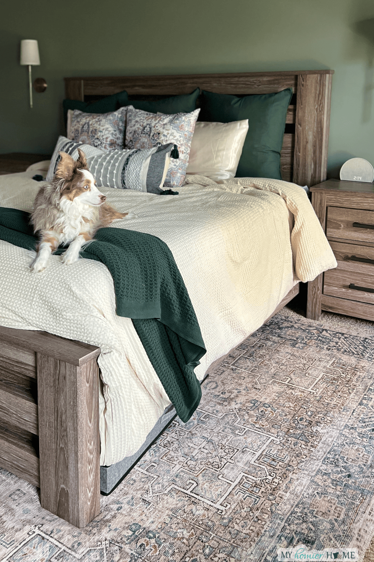 A dog sits on a neatly made bed with beige bedding, green accents, and multiple pillows; a nightstand is nearby, and a stylish rug under the king bed adds texture to the room.