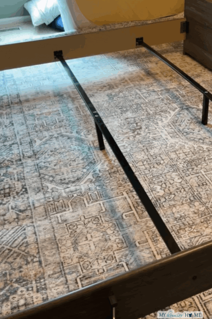 A partially assembled bed frame with metal support slats is positioned on a patterned rug under king bed in a bedroom.