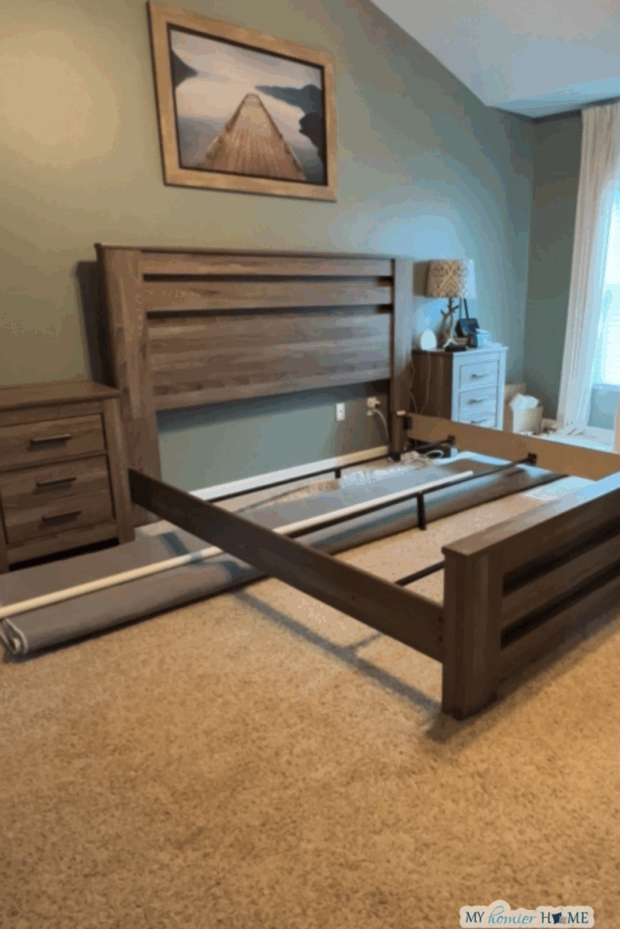 A partially assembled wooden bed frame stands on a carpeted floor with a rug under the king bed, beside a nightstand and lamp in a bedroom with sage green walls.
