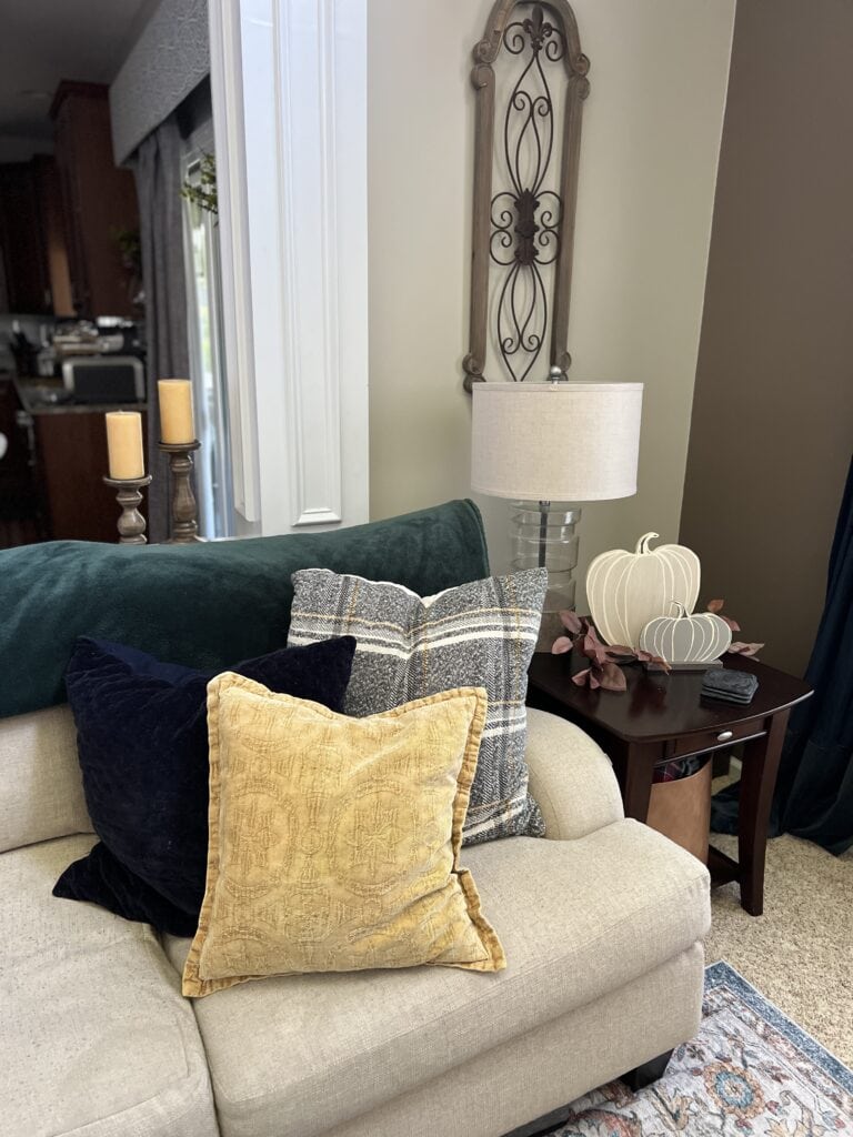 A beige couch with three decorative pillows sits next to a side table with a lamp, candles, and white pumpkin ornaments in a cozy living room, offering perfect fall home decor ideas.