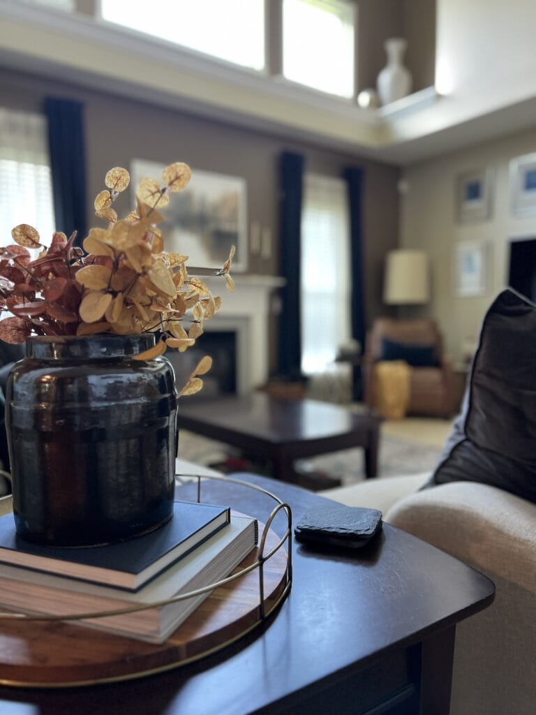 A black vase with dried flowers sits on books on a wooden table in a living room with a sofa, coffee table, and fireplace in the background, offering cozy inspiration for fall home decor ideas.