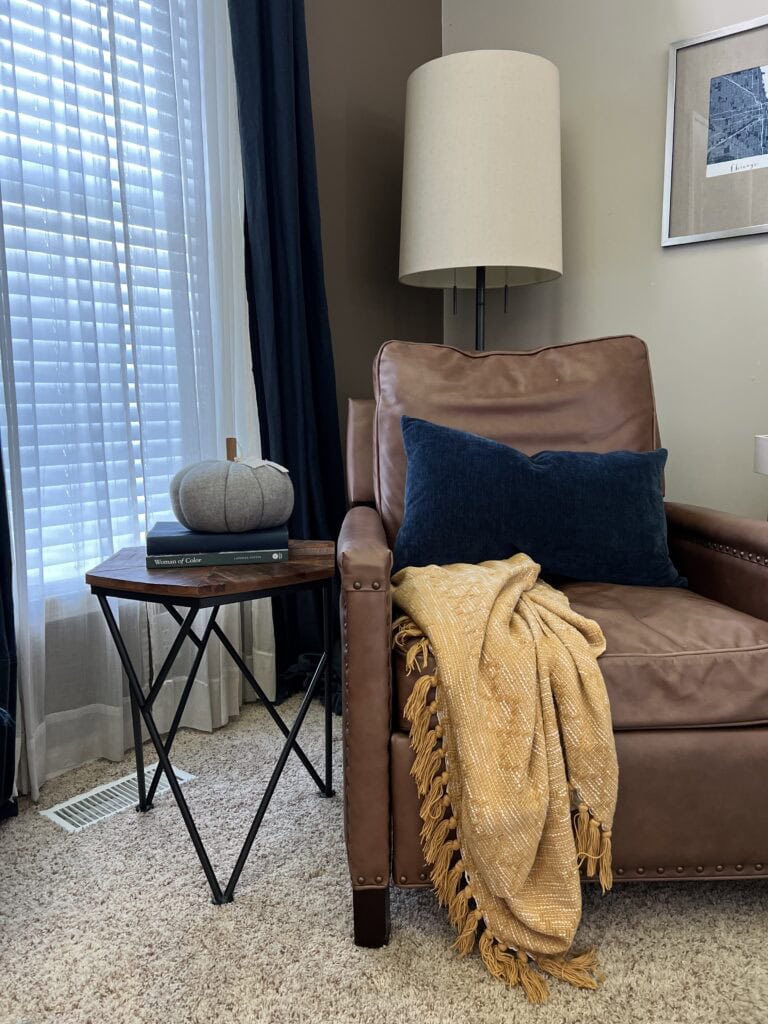 A brown leather armchair with a mustard throw and blue pillow sits by a window; a small table holds a fabric pumpkin and books, offering cozy fall home decor ideas, with a floor lamp and framed art on the wall.