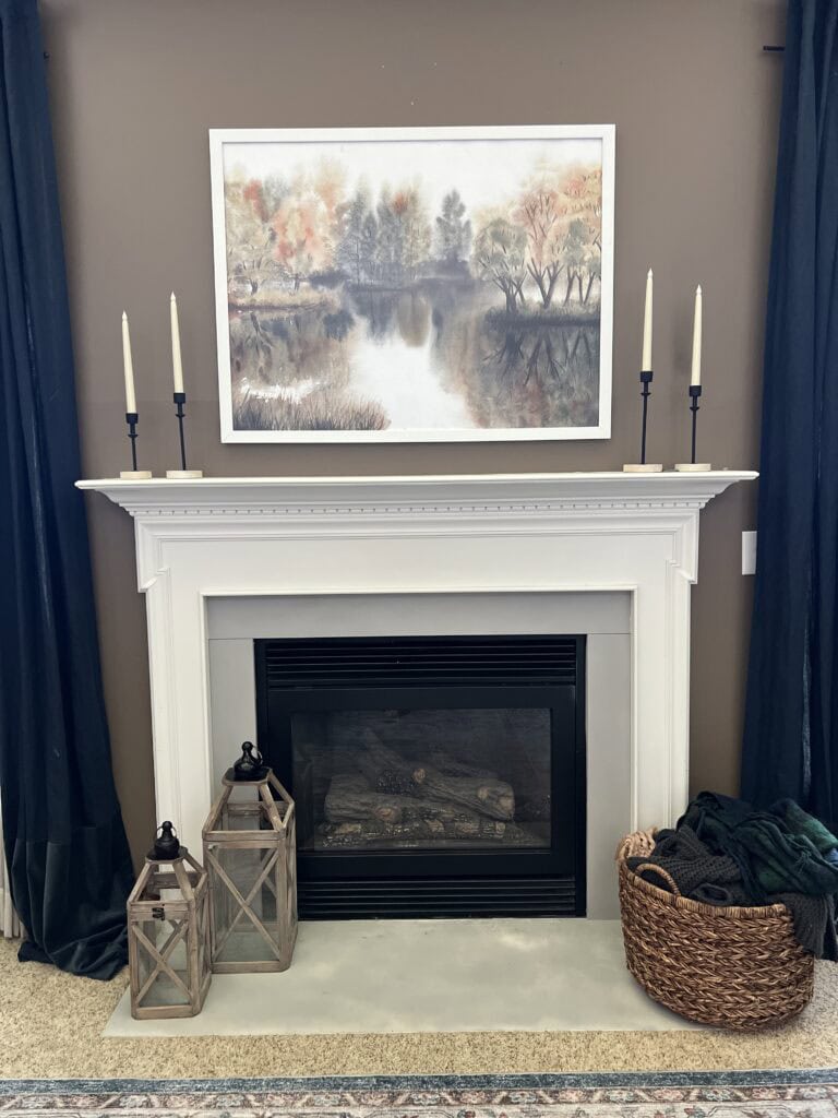 White fireplace with black interior, flanked by two lanterns and a woven basket—offering timeless inspiration for fall home decor ideas. Six candlesticks sit on the mantel beneath a framed landscape painting of trees by a lake, creating an inviting seasonal focal point.
