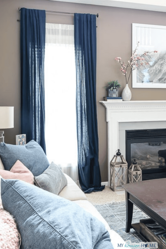 Living room with a white fireplace, blue curtains, a beige sofa with blue and pink pillows, and a coffee table. A large framed art piece and home decor are on the mantel, highlighting the inviting decorating style of this thoughtfully designed interior.