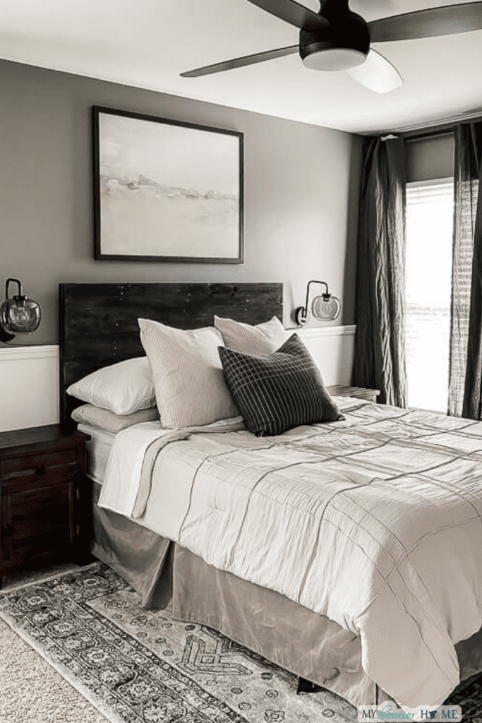 A neatly made bed with neutral bedding, dark wooden headboard, and modern ceiling fan showcases a stylish Room Refresh, complemented by abstract art and smart Small Room Design in this gray-toned bedroom.