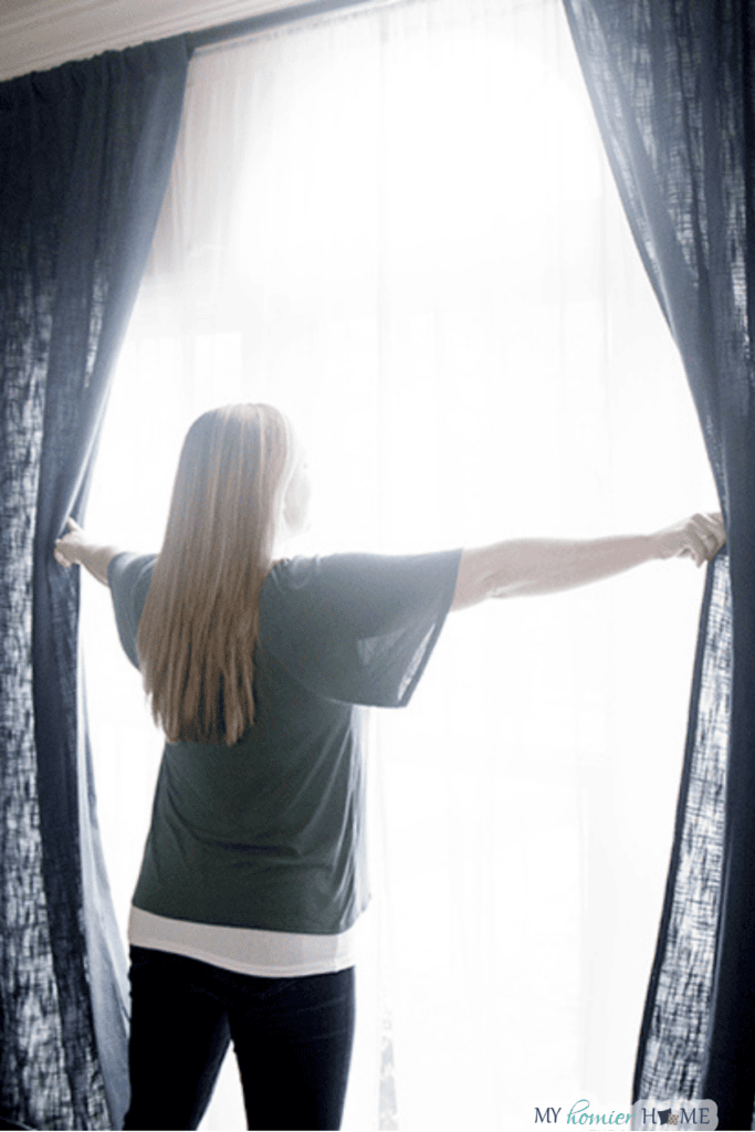 A woman with long hair stands facing a window, opening dark curtains to let in bright natural light as part of her weekly reset routine, bringing a sense of home calm to the space.
